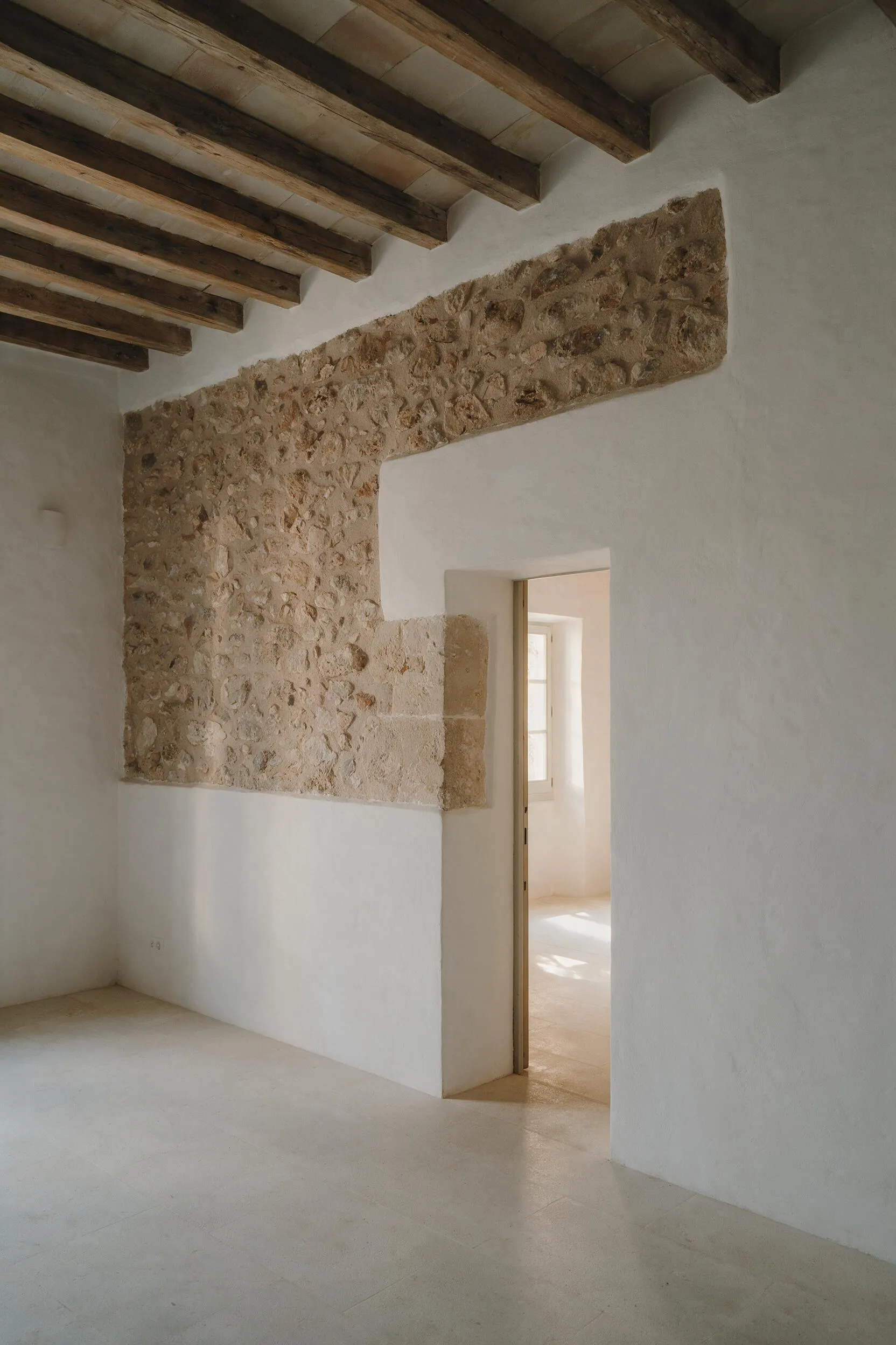 Interior de una finca mallorquina rehabilitada con muro de piedra visto, vigas de madera y paso a una segunda estancia